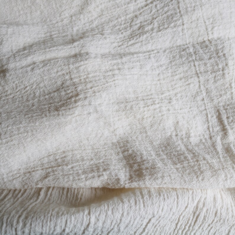 Puede incluir: Primer plano de una tela blanca texturizada con un aspecto suave y arrugado. La tela est&aacute; doblada por la mitad, creando una superficie lisa y uniforme.