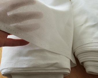 May include: Close-up of white fabric, possibly linen or cotton, with a hand holding a portion of the material. The fabric appears to be folded and stacked, with visible seams and a semi-sheer quality. The fabric is bright white.