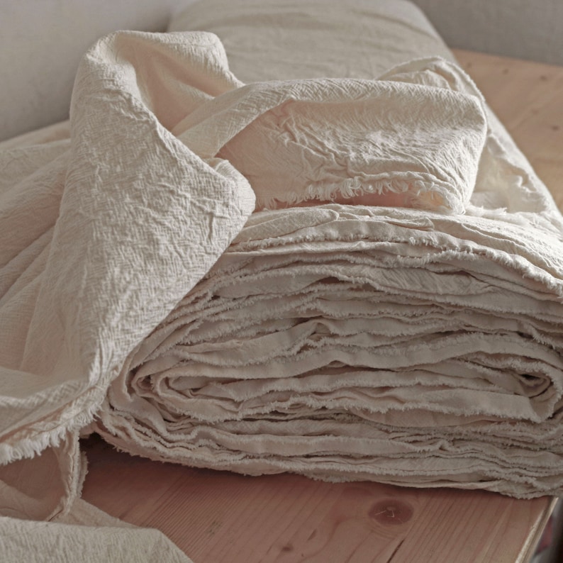 May include: A stack of off-white fabric with a frayed edge. The fabric is folded and piled on a wooden surface.