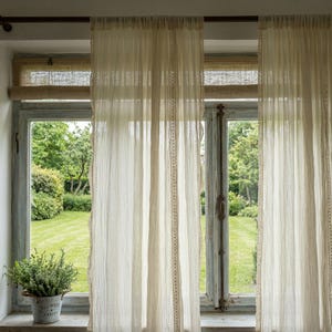 May include: Sheer, cream-colored curtains with delicate lace trim hang in front of a window. The window frame is a weathered blue, and a small potted plant sits on the windowsill. The curtains filter the sunlight, creating a soft, diffused glow.