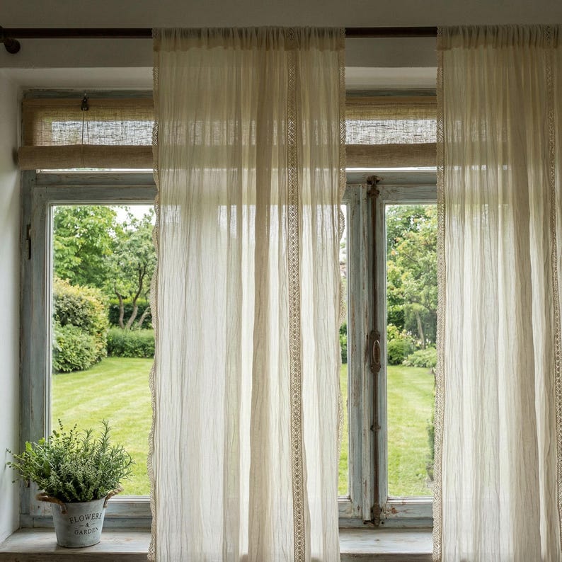 May include: Sheer, cream-colored curtains with delicate lace trim hang in front of a window. The window frame is a weathered blue, and a small potted plant sits on the windowsill. The curtains filter the sunlight, creating a soft, diffused glow.
