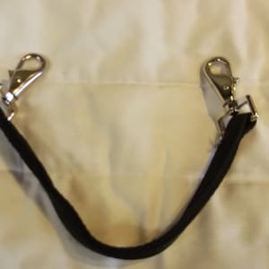 Puede incluir: Una correa negra con dos cierres giratorios de metal plateado. Los cierres están sujetos a la correa, que está curvada en forma de U. El fondo es de tela blanca.