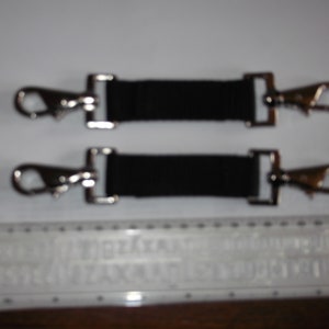 Puede incluir: Dos correas de cincha negras con mosquetones giratorios plateados. Cada correa tiene un rectángulo de metal en cada extremo. Los ganchos están sujetos a los rectángulos de metal. Longitud aproximada de 10 cm.