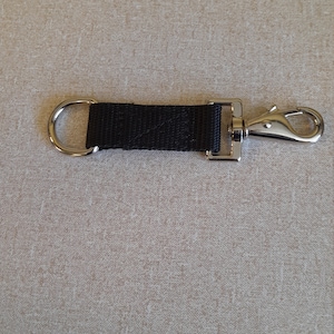 May include: A black nylon strap with a silver metal swivel clasp and a D-ring. The strap is attached to the clasp with a metal rectangle. The item is laying on a beige surface.