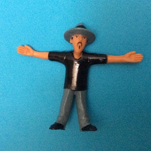 May include: A plastic figurine of a person wearing a blue hat and black shirt with gray pants. The figurine has a mustache and is standing with arms outstretched.