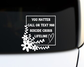 May include: White decal with the text "YOU MATTER CALL OR TEXT 988 SUICIDE CRISIS LIFELINE" inside a white square, with floral and leaf accents. The decal is on the rear window of a white vehicle.