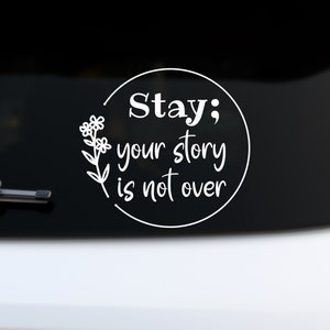 May include: White vinyl decal with a black circle and the text "Stay; your story is not over" with a small flower design to the left of the text.