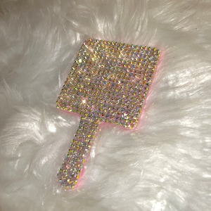 May include: A pink hand mirror covered in iridescent rhinestones. The mirror has a square shape and a handle.