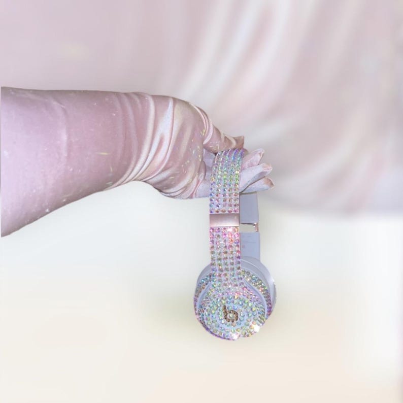 May include: A pair of light blue headphones with iridescent rhinestones covering the entire surface. The headphones are held by a hand wearing a white glove.