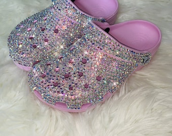 May include: A pair of pink clogs covered in iridescent rhinestones. The clogs are on a white fluffy surface.