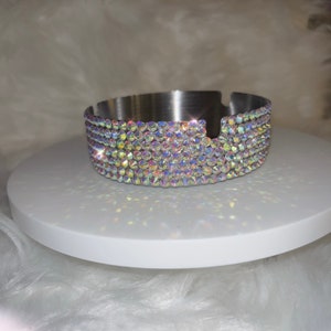 May include: A silver ashtray with a decorative iridescent crystal trim. The ashtray is round and has a small indentation for cigarettes.