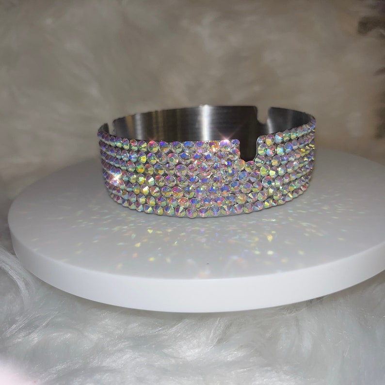 May include: A silver ashtray with a decorative iridescent crystal trim. The ashtray is round and has a small indentation for cigarettes.