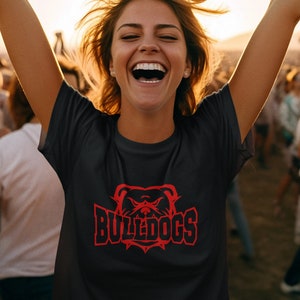 Op de afbeelding: Een zwart T-shirt met een rode bulldog-afbeelding en de tekst "BULLDOGS" gedrukt op de voorkant. De persoon die het shirt draagt, glimlacht en heeft zijn armen in de lucht gestoken.