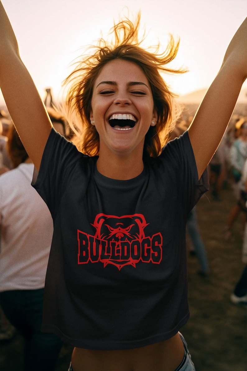Op de afbeelding: Een zwart T-shirt met een rode bulldog-afbeelding en de tekst "BULLDOGS" gedrukt op de voorkant. De persoon die het shirt draagt, glimlacht en heeft zijn armen in de lucht gestoken.
