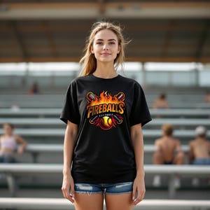 May include: Black t-shirt featuring the text "Fireballs Softball" in a fiery design. The graphic includes a softball, bats, and flames. The shirt is paired with denim shorts.