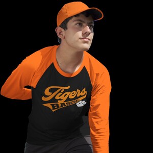 May include: A black and orange baseball jersey with the text "Tigers Baseball" in a stylized font. The jersey has a white paw print logo and is worn by a person wearing an orange baseball cap.