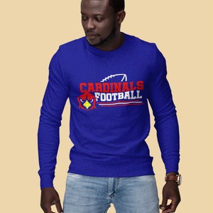 Puede incluir: Una camiseta de manga larga azul real con un dise&ntilde;o gr&aacute;fico blanco y rojo que dice "Cardinals Football" con un gr&aacute;fico de p&aacute;jaro cardenal rojo y un gr&aacute;fico de f&uacute;tbol blanco.