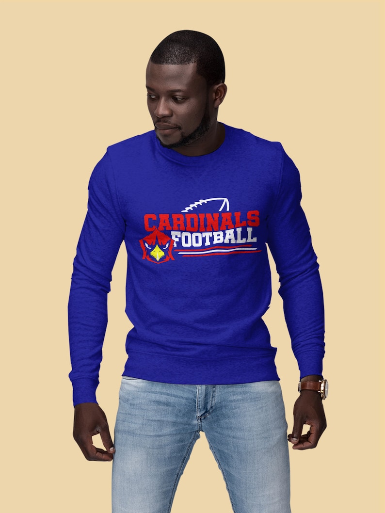 Puede incluir: Una camiseta de manga larga azul real con un dise&ntilde;o gr&aacute;fico blanco y rojo que dice "Cardinals Football" con un gr&aacute;fico de p&aacute;jaro cardenal rojo y un gr&aacute;fico de f&uacute;tbol blanco.