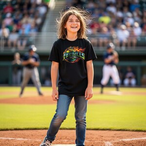 May include: A person wearing a black t-shirt with the words "Fireballs Softball" in a colorful design. The person is wearing blue jeans and standing on a baseball field. The background shows a baseball game in progress.