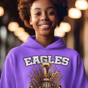 K&ouml;nnte beinhalten: Lilafarbener Hoodie mit einer wei&szlig;en Grafik einer Gruppe von Musikinstrumenten, darunter eine Trompete, eine Posaune und eine Klarinette. Die Grafik enth&auml;lt auch den Text "EAGLES BAND".