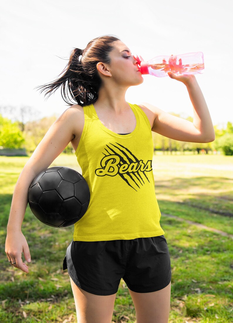 May include: A woman wearing a yellow tank top with the word "Bears" in black lettering and a black claw mark design. She is holding a black soccer ball and drinking from a pink water bottle.