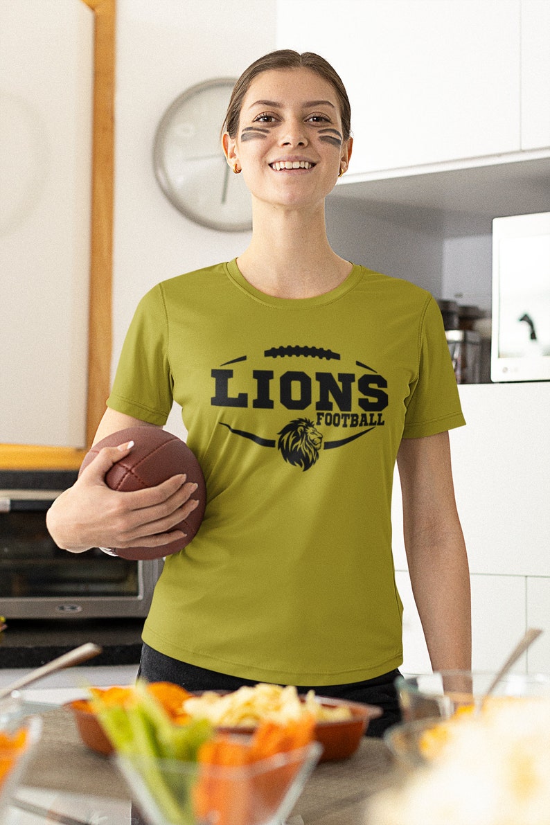 May include: A woman wearing a green t-shirt with a black lion graphic and the text "Lions Football". She is holding a brown football.