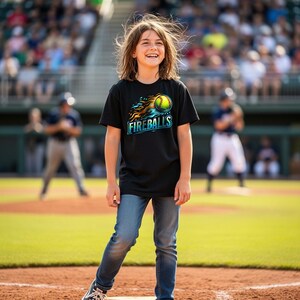 Puede incluir: Una persona con una camiseta negra con un gr&aacute;fico de b&eacute;isbol que dice "Fireballs" en azul y una pelota de b&eacute;isbol con llamas. La persona lleva vaqueros azules y est&aacute; de pie en un campo de b&eacute;isbol.