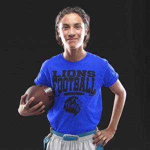 May include: A person wearing a royal blue t-shirt with the words "LIONS FOOTBALL" and a lion graphic. The person is holding a brown football. The background is black.