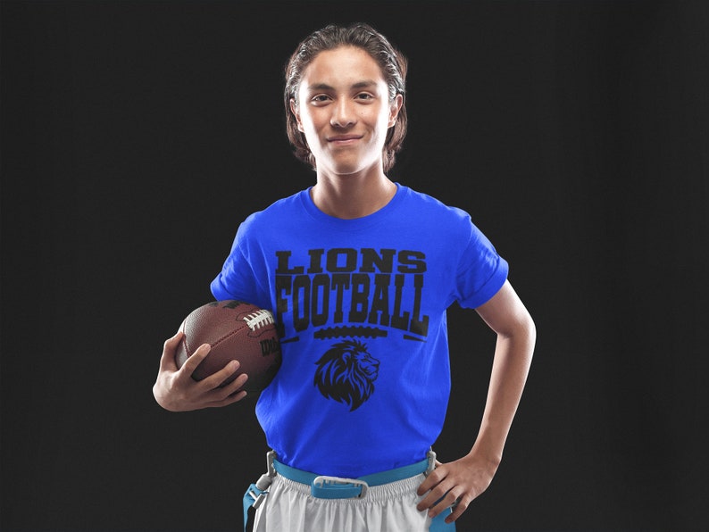 May include: A person wearing a royal blue t-shirt with the words "LIONS FOOTBALL" and a lion graphic. The person is holding a brown football. The background is black.