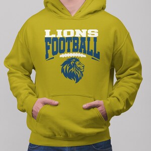 May include: Mustard yellow hoodie with a front pocket and a hood. The front of the hoodie features the text "LIONS FOOTBALL" in white and blue, along with a blue lion logo. The person is wearing blue jeans.