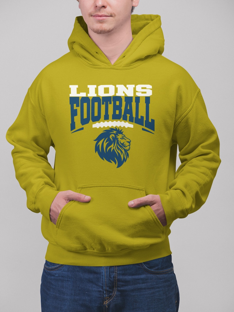 May include: Mustard yellow hoodie with a front pocket and a hood. The front of the hoodie features the text "LIONS FOOTBALL" in white and blue, along with a blue lion logo. The person is wearing blue jeans.