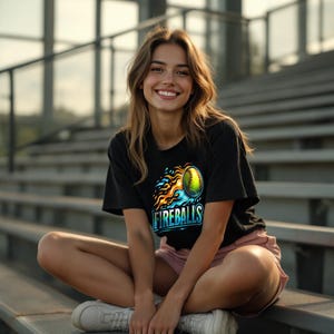 Puede incluir: Camiseta negra con un gr&aacute;fico de softbol y la palabra "FIREBALLS" en blanco y azul. El dise&ntilde;o de softbol presenta una pelota amarilla con llamas azules y naranjas. La persona lleva pantalones cortos rosas y zapatillas blancas.