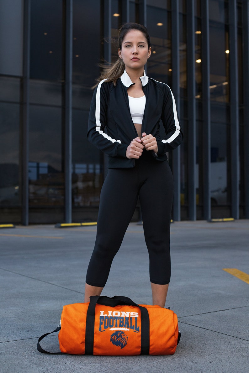 May include: A woman wearing a black athletic jacket with white stripes, a white sports bra, and black capri leggings. An orange duffel bag with "LIONS FOOTBALL" text and a lion logo is at her feet.