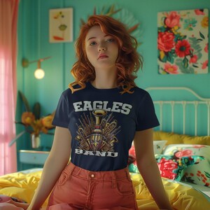 K&ouml;nnte beinhalten: Eine Frau tr&auml;gt ein marineblaues T-Shirt mit einem Motiv von Musikinstrumenten und dem Text "Eagles Band". Sie tr&auml;gt orangefarbene Shorts und sitzt auf einem gelben Bett.