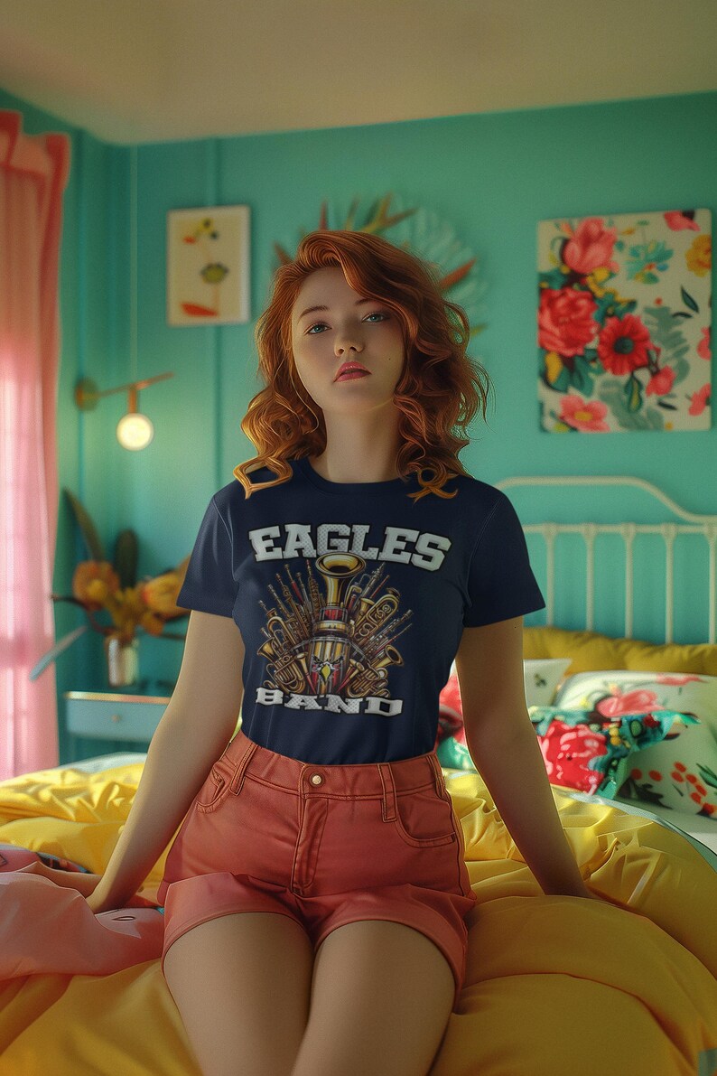 K&ouml;nnte beinhalten: Eine Frau tr&auml;gt ein marineblaues T-Shirt mit einem Motiv von Musikinstrumenten und dem Text "Eagles Band". Sie tr&auml;gt orangefarbene Shorts und sitzt auf einem gelben Bett.