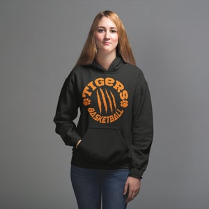 Puede incluir: Sudadera con capucha negra con un gr&aacute;fico circular naranja que dice "Tigers Basketball" con huellas de patas y marcas de garras.