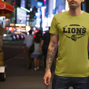 May include: A man wearing a yellow t-shirt with a black graphic of a football with the word "LIONS" and "FOOTBALL" underneath. The man has tattoos on his arms.