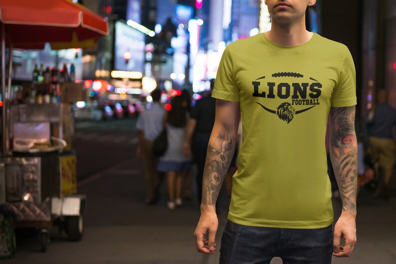 May include: A man wearing a yellow t-shirt with a black graphic of a football with the word "LIONS" and "FOOTBALL" underneath. The man has tattoos on his arms.