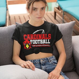 Puede incluir: Una camiseta negra con un gr&aacute;fico rojo y blanco que dice "CARDINALS FOOTBALL" con una ilustraci&oacute;n de un p&aacute;jaro cardenal rojo. La camiseta est&aacute; anudada a la cintura y la persona que la lleva est&aacute; sentada en un sof&aacute; gris.