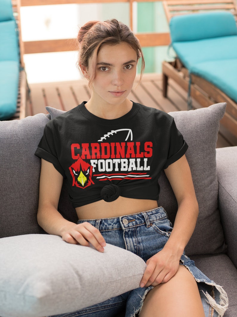 Puede incluir: Una camiseta negra con un gr&aacute;fico rojo y blanco que dice "CARDINALS FOOTBALL" con una ilustraci&oacute;n de un p&aacute;jaro cardenal rojo. La camiseta est&aacute; anudada a la cintura y la persona que la lleva est&aacute; sentada en un sof&aacute; gris.