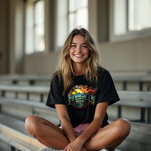 May include: A black t-shirt featuring a softball design with the text "Fireballs Softball" in a colorful, dynamic graphic. The shirt is worn by a person sitting with legs crossed, smiling. The person is wearing pink shorts.