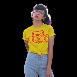 Op de afbeelding: Een geel T-shirt met een rode bulldog-afbeelding en de tekst "BULLDOGS" in rood. De persoon die het shirt draagt, draagt een blauwe jeans en heeft paars haar.