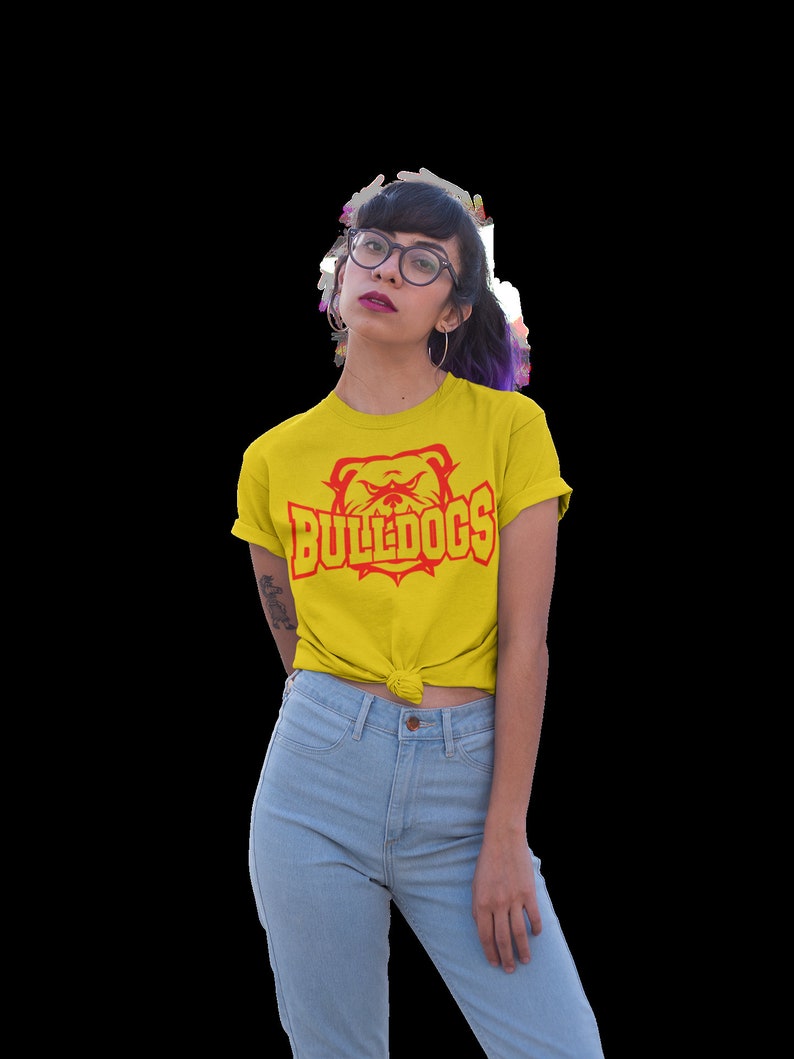 Op de afbeelding: Een geel T-shirt met een rode bulldog-afbeelding en de tekst "BULLDOGS" in rood. De persoon die het shirt draagt, draagt een blauwe jeans en heeft paars haar.