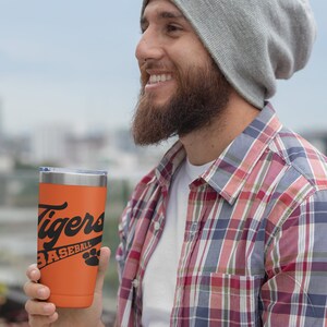 May include: A person wearing a gray beanie and a red and blue plaid shirt is holding an orange travel mug with black text that reads "Tigers Baseball" and a paw print.