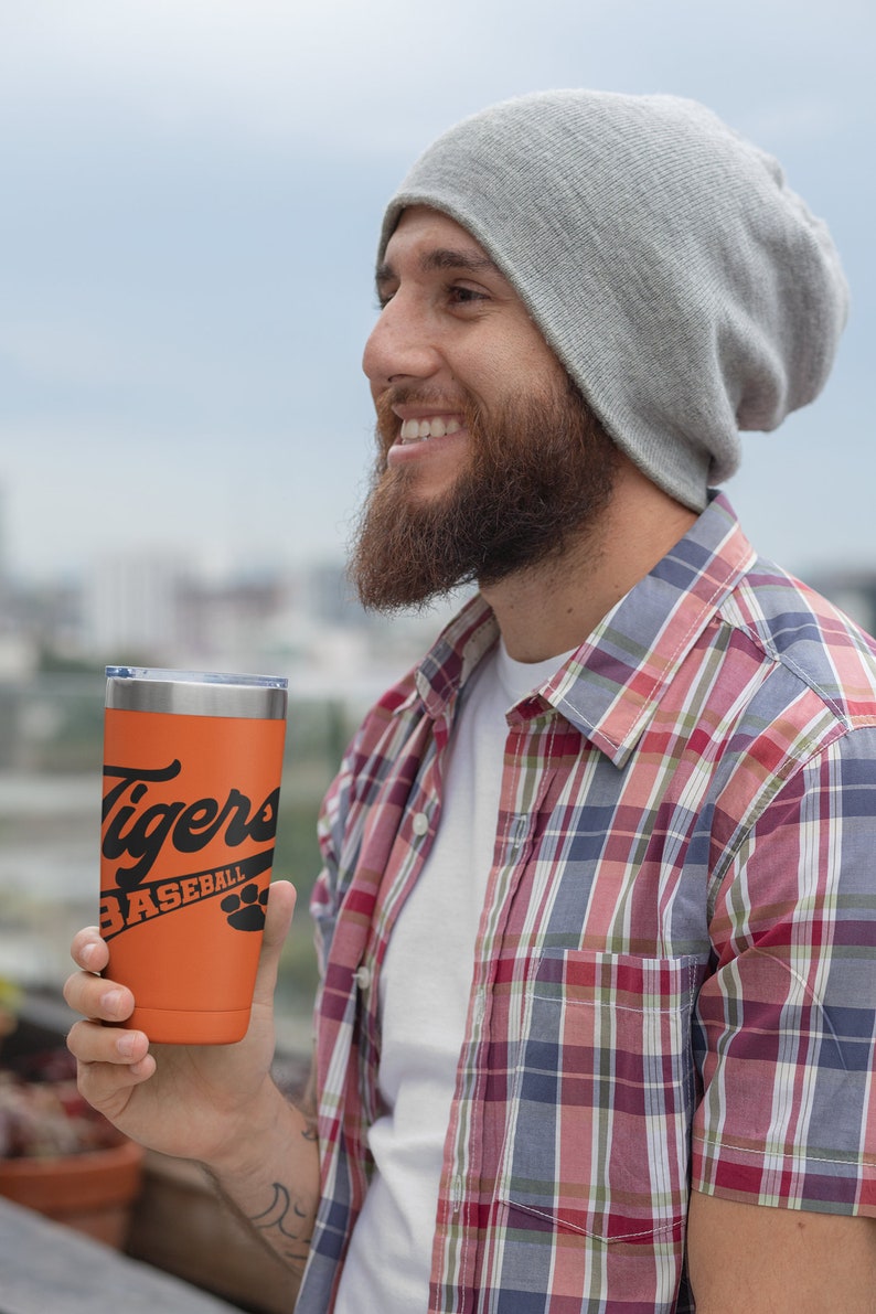 May include: A person wearing a gray beanie and a red and blue plaid shirt is holding an orange travel mug with black text that reads "Tigers Baseball" and a paw print.