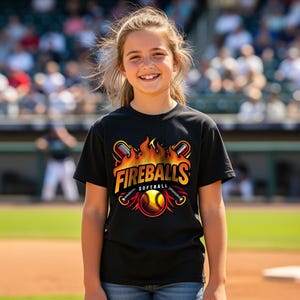 May include: A black t-shirt with the words "Fireballs Softball" in yellow and orange, featuring a softball design with flames and bats. The shirt is worn by a person standing on a baseball field.