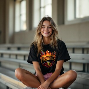 May include: Black t-shirt with a softball graphic. The design includes the word "Fireballs" in gold with flames, bats, and a softball. The shirt is worn by a person.