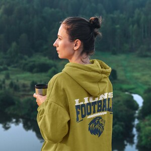 May include: Mustard-colored hoodie with "LIONS FOOTBALL" printed on the back in navy blue and white, along with a lion graphic. The hoodie is worn by a person holding a travel mug, with a scenic background of a forest and water.