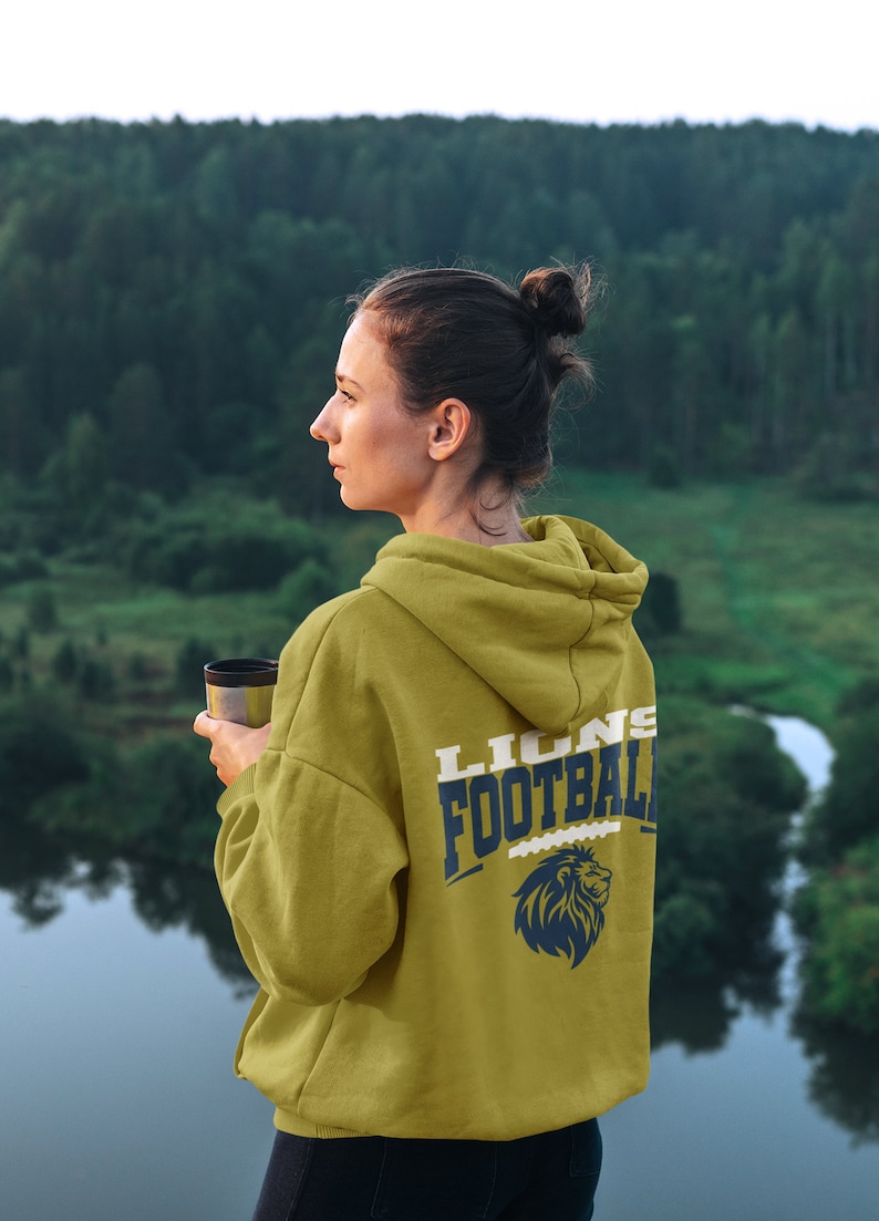 May include: Mustard-colored hoodie with "LIONS FOOTBALL" printed on the back in navy blue and white, along with a lion graphic. The hoodie is worn by a person holding a travel mug, with a scenic background of a forest and water.
