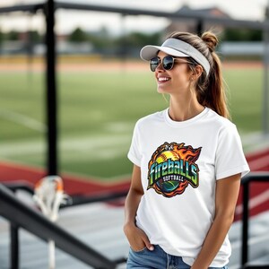 May include: White t-shirt with a colorful graphic of a softball engulfed in flames, with the text "Fireballs Softball". The shirt is short-sleeved and has a crew neck. The design features shades of yellow, orange, green, and blue.